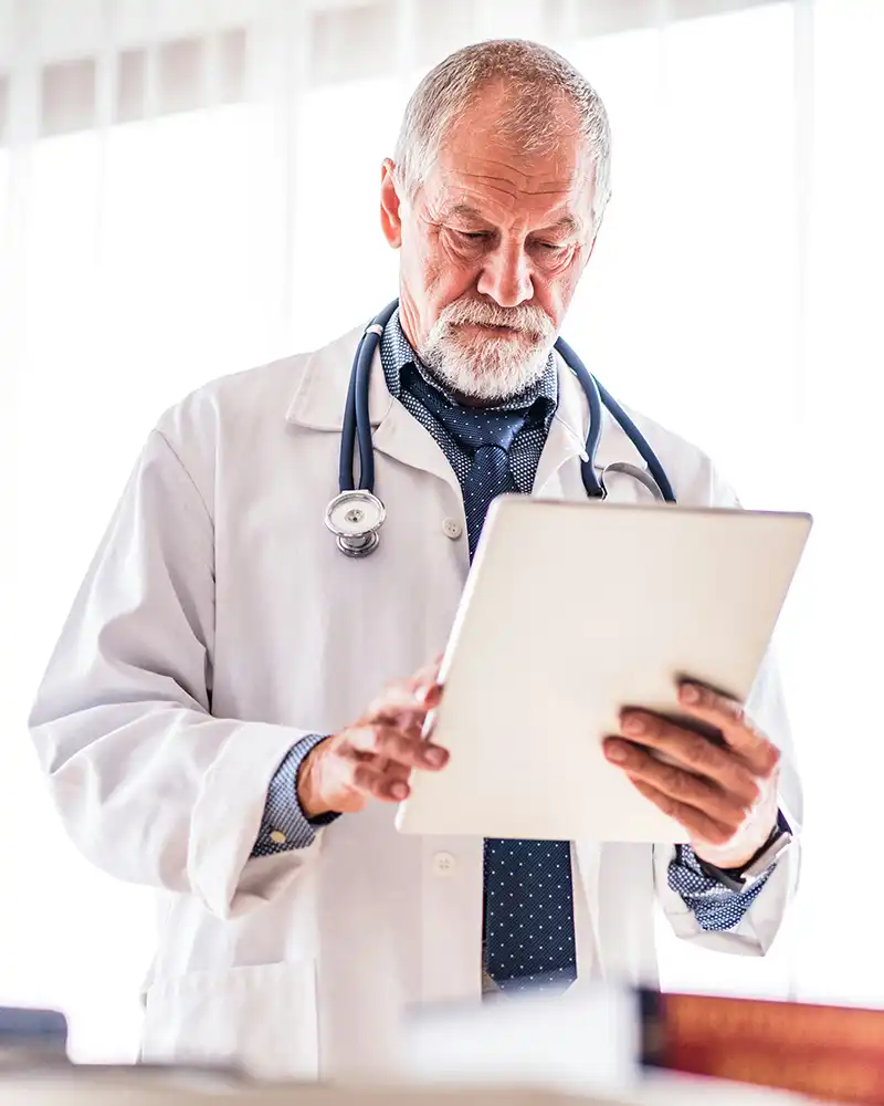 An older male doctor with a stethoscope around his neck reading a patient's chart.